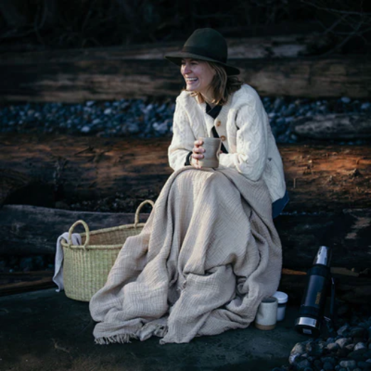 Home | Elizabeth Homestead (Model sitting on a chair covered with a comfy blanket holding a mug wearing a knit sweater with a wicker basket next to her.)