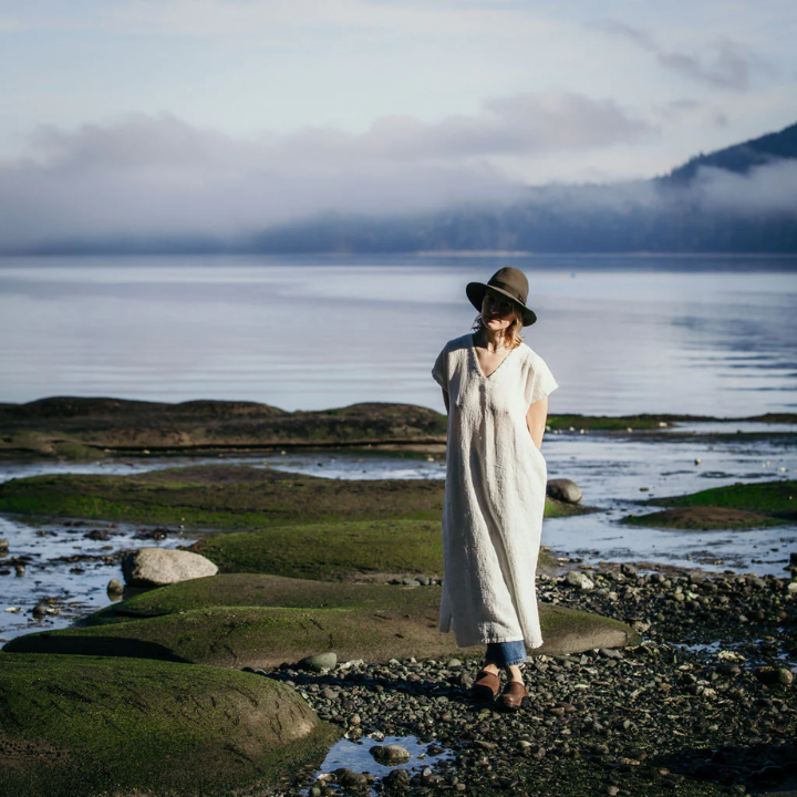 Moss Grey | Elizabeth Homestead (Model standing on the shore of a body of water wearing a fedora hat and Moss Grey long linen dress)